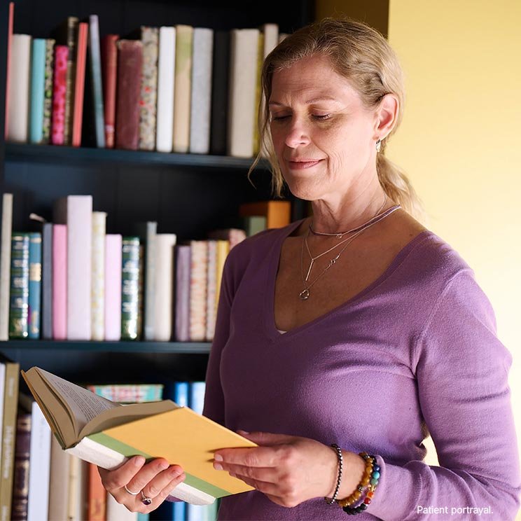 A woman reading a book stands next to a bookcase.